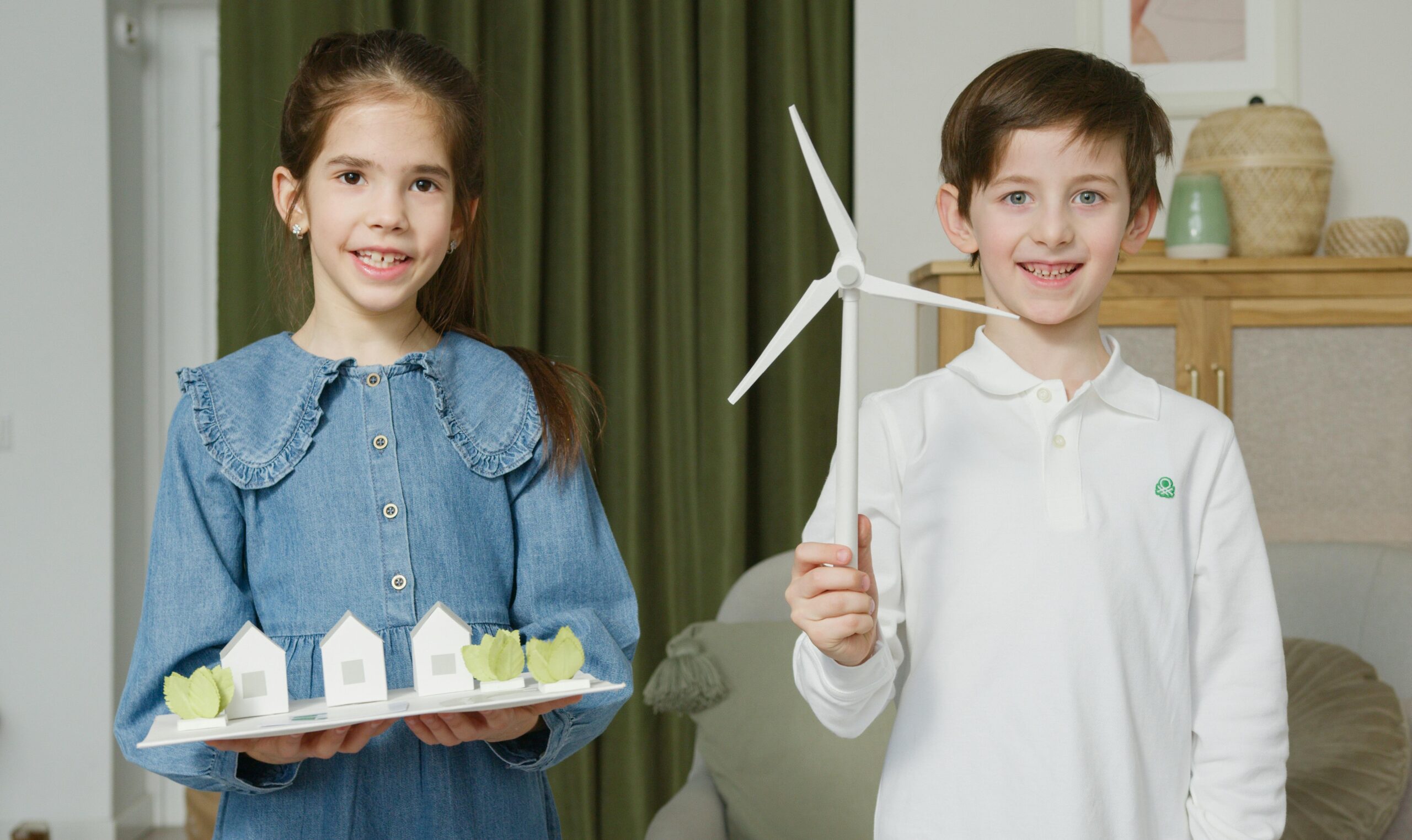 Two children presenting a wind turbine model and eco-friendly homes indoors for learning.